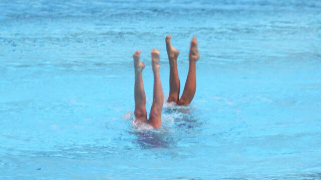 Two artistic swimmers performing synchronized leg movements during a duet routine in a swimming pool