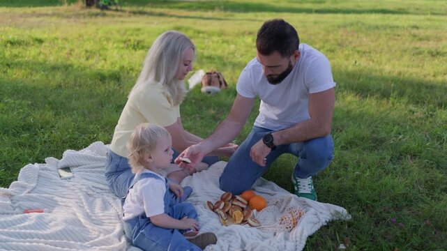 A young family with a toddler enjoys a picnic on a sunny day. The loving father carefully feeds his baby son bread while sitting on a blanket in a green park. A heartwarming family moment.