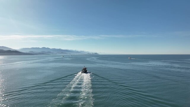 Drone shot capturing a fishing vessel moving through the calm blue waters of the Black Sea. The boat leaves a distinct white wake behind it as it travels toward the distant skyline of Batumi city.