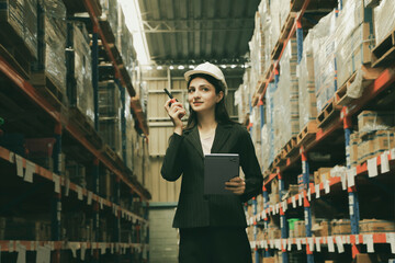 Fototapeta premium Professional female worker wearing hard hat works in industrial distribution center in factory warehouse using tablet digital and walkie talkie communication to check logistics transportation.