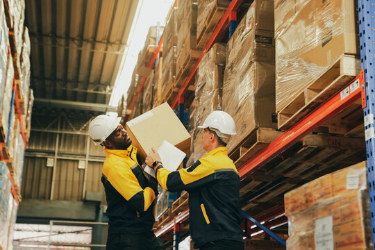 Two male warehouse workers are helping each other lift heavy cardboard boxes onto shelves. The two workers support each other to complete the task : Two workers are carrying the boxes manually without