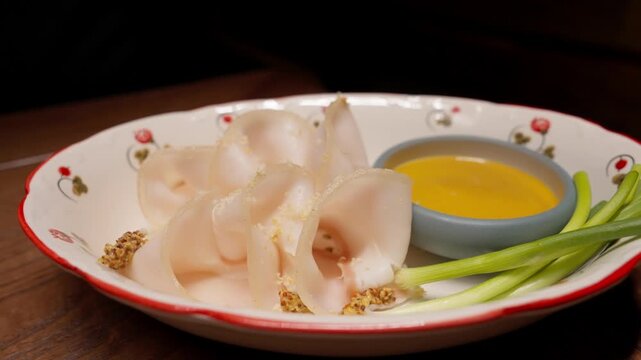 Thinly sliced cured pork fat arranged on a decorative plate with mustard sauce and fresh green onions, presented as a rustic appetizer on a wooden table