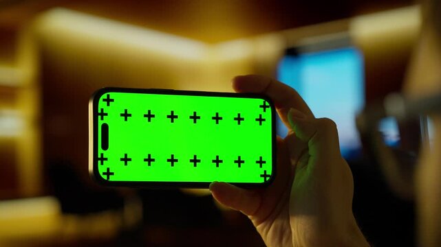 Close-up hand holding smartphone with vivid green chroma key screen and tracking markers in warm blurred indoor environment for digital compositing