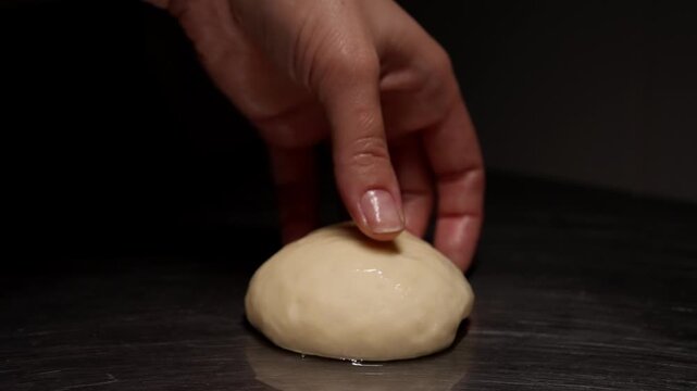 Close-up sequence of soft bread dough resting and being gently pressed by a hand on a dark metal countertop in a minimalist kitchen baking preparation scene