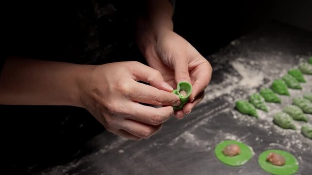 Close up of skilled hands shaping vibrant green dumplings with savory filling on a floured work surface, highlighting meticulous homemade cooking craftsmanship
