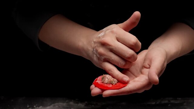 Closeup of skillful hands shaping a delicate red dumpling by filling and folding dough against a dark background in a detailed culinary preparation process