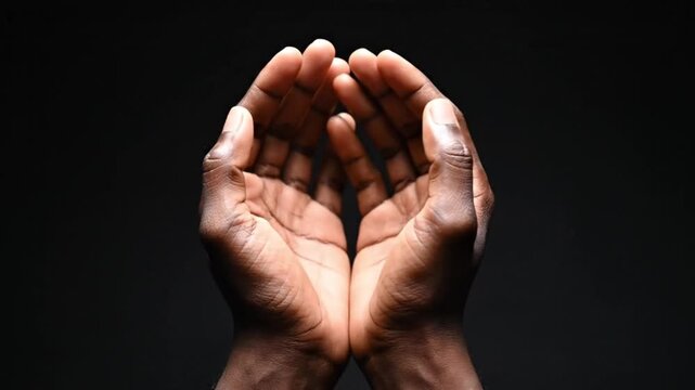 A close-up shows two dark-skinned hands, gently cupped and open against a stark black background. This profound, expressive gesture conveys universal themes of offering, receiving, prayer, hope, and c