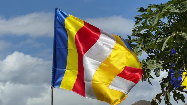 A colorful Buddhist flag flutters actively in the wind near green trees under a cloudy sky in a tranquil environment. Cultural heritage.