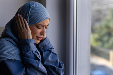 Woman afraid as hearing explosions outside her window in busy city, standing covering ears and...