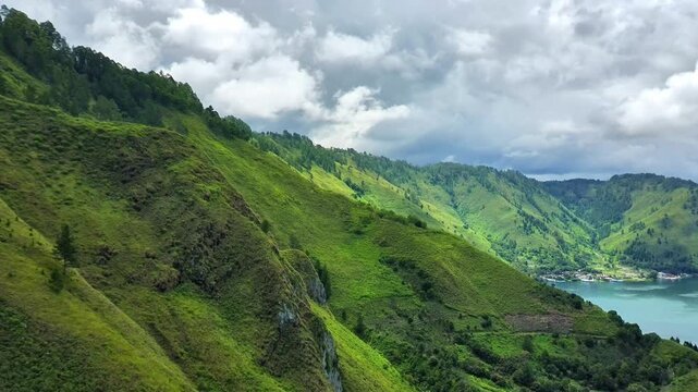 4K aerial drone footage flying near a green hillside along Lake Toba, showing lush vegetation, pine trees, and rolling hills with a glimpse of calm lake water under cloudy daylight