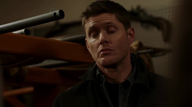 Man contemplates seriousness while near weapons and firearms collection indoors, dramatic portrayal, male.