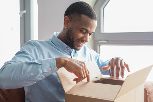 A smiling African American man sits on sofa at home unpacking a parcel, looking happily inside a cardboard box after receiving an online order and enjoying a reliable delivery service