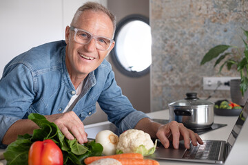 Fototapeta premium Smiling mature man searching for new recipes on his laptop while cooking healthy food at home in a modern kitchen, preparing vegetables. Concept of nutrition, active lifestyle, and healthy eating
