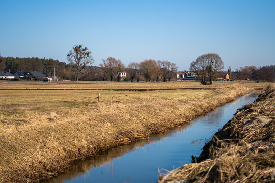 Narrow irrigation canal flowing through agricultural fields, rural landscape with river and dry grass on sunny day, blue sky over countryside and trees in early spring, water retention.