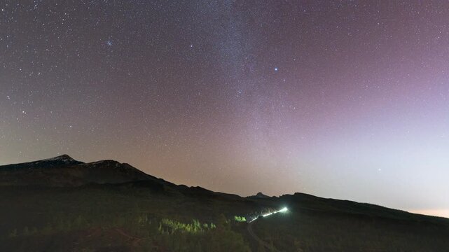 4K timelapse of the Milky Way arching over Mount Teide volcano in Tenerife at night. Stars rotate above volcanic landscape, faint car headlights trace roads in the valley below