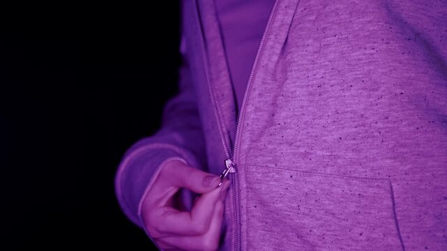A close-up view of a female hand pulling down the metal zipper of a grey heather hoodie against a black background. Concept of casual street style, night life, and modern fashion in ultraviolet light