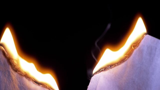 Edges of white paper slowly burn with bright glowing flames and smoke on a black background. Concept of destruction, eliminating documents, and creating dark abstract transitions. Slow motion.