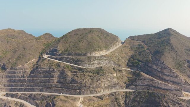 Aerial view of a winding asphalt road carving through steep rocky mountains with cars traversing switchbacks near the coast in Iran.