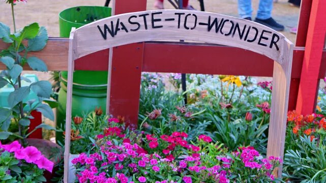 A WASTE-TO-WONDER sign arches over a colorful flower bed. Bright pink and orange blossoms grow within a rustic garden display, with visitors' legs visible in the background.