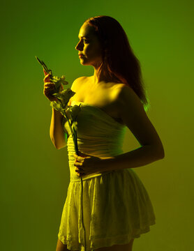 Close up portrait of female model with red hair and white dress, holding flowers in side profile.  Isolated figure with colourful creative studio lighting with golden glowing silhouette and green back