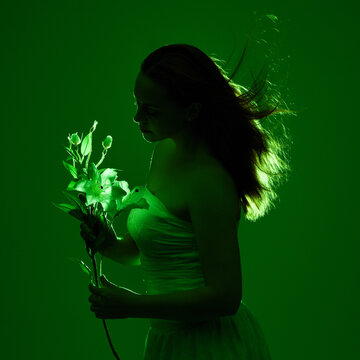 Close up portrait of female model with red hair and white dress, holding flowers in side profile.  Isolated figure with colourful creative studio lighting with golden glowing silhouette and green back