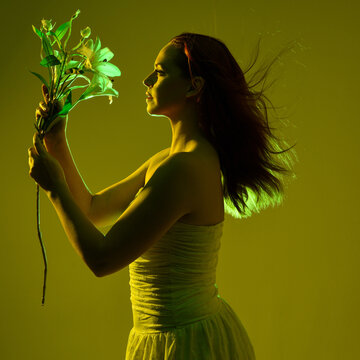 Close up portrait of female model with red hair and white dress, holding flowers in side profile.  Isolated figure with colourful creative studio lighting with golden glowing silhouette and green back