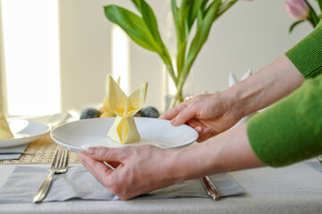 Woman serving table for Easter holiday family dinner, hands holding plate with yellow napkin rabbit decoration, fresh spring table setting with flowers