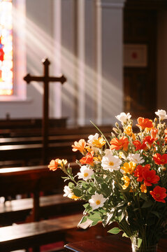Vertical shot of spring flowers in a church with pews and a cross in the backdrop. Sunlight streaming through window