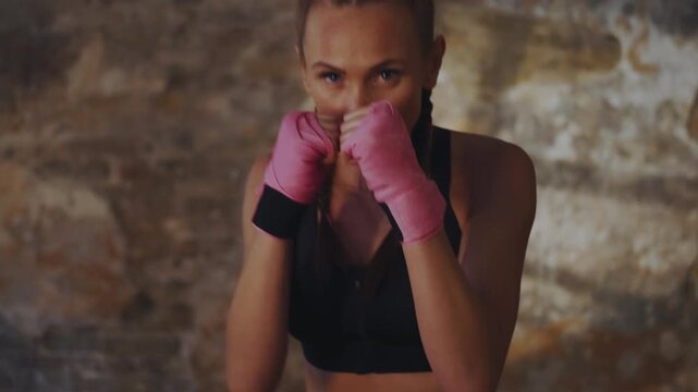 Determined woman with braids and pink boxing wraps holds a focused fighting stance during a shadowboxing session in a gritty indoor setting
