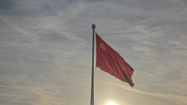 The historical flag of the USSR, with its iconic hammer and sickle and red star, flutters proudly on a flagpole against a clear sky.