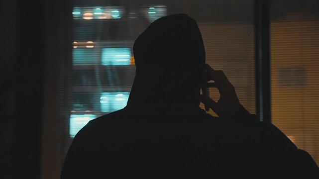 Mysterious man with back to camera standing by apartment window talking on phone. Spy or hacker undercover operation or illegal criminal activity at night speaking on mobile device making shady deal