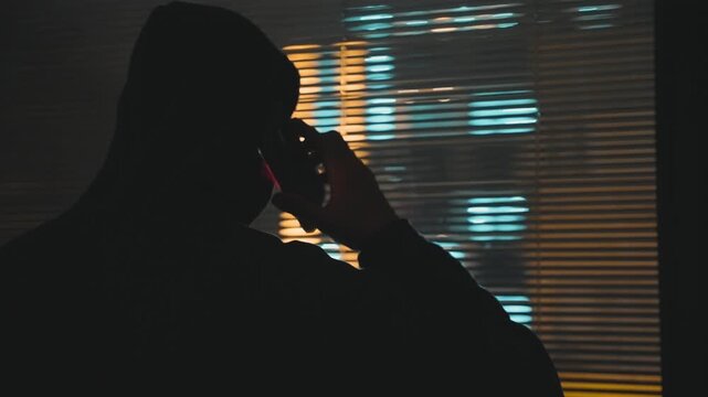Hooded man stands by apartment window talking on mobile phone at night. Suspicious and mysterious person with black hoodie spy agent or hacker holding smartphone making call conversation in dark room