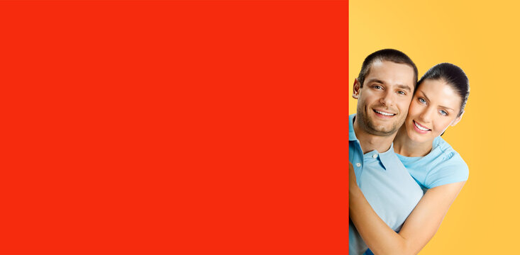 Portrait image - happy smile embracing hugging cute joyful playful young couple man woman peep from red empty sign board billboard with free ad space, isolated yellow background. Mock up mockup banner