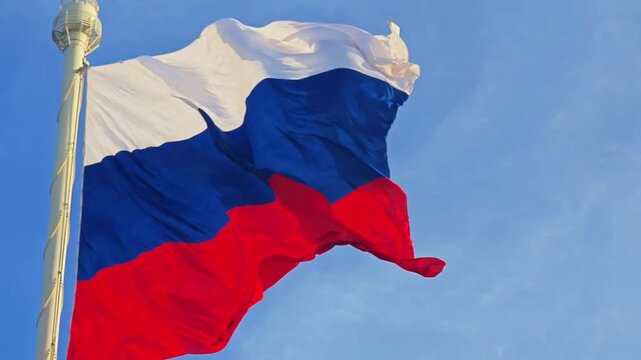 A flagpole with the Russian Federation tricolor flag waving dynamically in the wind against a clear sky background. Patriotic national symbol.