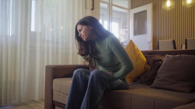 Stomach cramps, hands and woman on sofa in home with menstruation, periods or constipation. Tummy pain, sick of female person rubbing belly for IBS, endometriosis or virus in living room