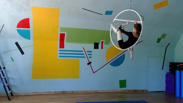 Nine year old girl practices aerial gymnastics tricks on aerial hoop in sports school with safety harness and elastic bandages on legs, slow motion, strength flexibility training