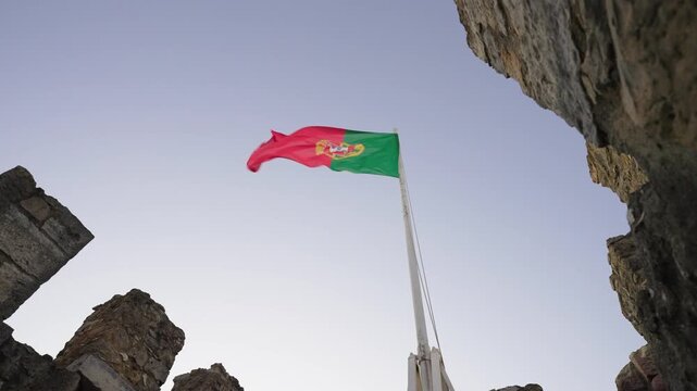 The national flag of Portugal waving from a flagpole between medieval stone battlements at Castelo de S&atilde;o Jorge, Lisbon, Portugal.