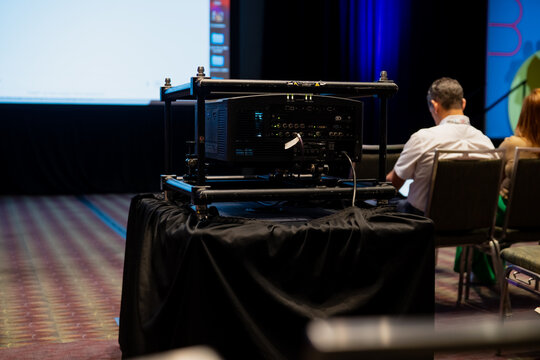 Audio visual control rack on a draped table in a conference room with projection screen and seated attendees, capturing live event production and meeting technology