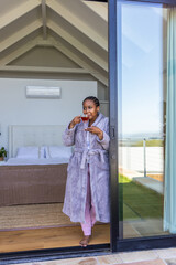 Naklejka premium African American female standing in bedroom doorway in bathrobe, sipping teacup near sliding door