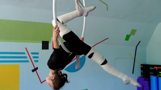 Nine year old girl practices aerial gymnastics tricks on aerial hoop in sports school with safety harness and elastic bandages on legs, slow motion, strength flexibility training