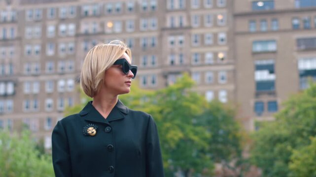 Caucasian woman wearing sunglasses and coat with a lovely brooch. Low angle view at the female in classic outfit outdoors in the blurred backdrop.