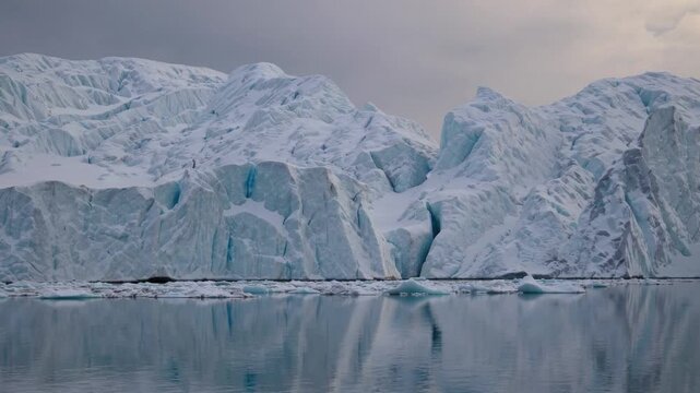 A vast glacier and icebergs reflected in calm water under a cloudy, overcast sky, depicting a serene polar landscape.