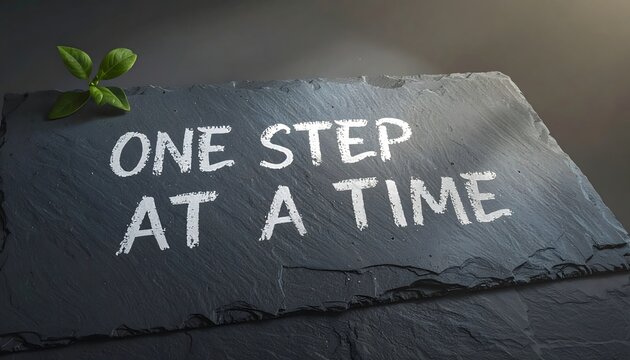 Motivational quote one step at a time written in white chalk on a textured dark slate stone background with green leaves