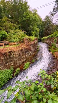 Mountain stream flowing through stone walls and green woodland area. View of a small river channel with stone embankments surrounded by green trees and wooden construction in Jim Thorpe.