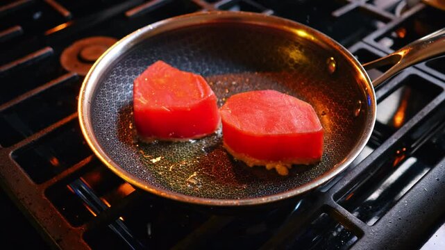 Two fresh tuna steaks searing in frying pan. Close up of two red tuna fillets frying in oil inside a stainless steel pan with hexagonal texture on gas stove.