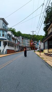 Woman in black dress walking on street in Jim Thorpe Pennsylvania. Woman walks down a paved road lined with colorful historic houses and American flags under an overcast sky.
