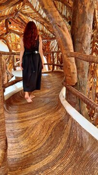 Redheaded woman in black dress walking on wooden bridge. Back view of woman with long red hair walking barefoot on a curved organic wooden pathway.