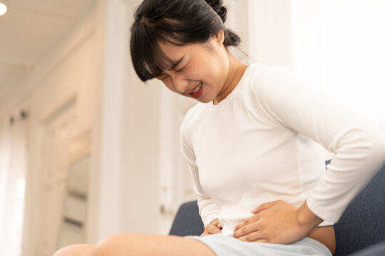 Asian woman sitting on sofa holding stomach with painful expression, suffering from abdominal pain at home, concept of stomachache, illness, health problem. woman, stomachache, abdominal, pain.
