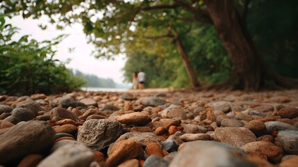 A couple walks away on a pebble beach beside tranquil water with lush trees and soft bokeh in the background
