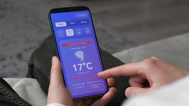 Close-up of hands holding smartphone displaying a red hurricane warning alert. User checking severe weather forecast, wind speed, and temperature on a modern mobile app interface during a storm